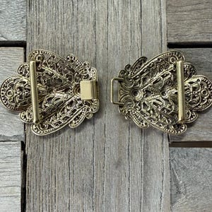May include: Two-piece gold-colored filigree buckle set. Each piece has an ornate, openwork design with a bar and a clasp. The buckle is likely for a belt or other garment, and is displayed on a wooden surface.