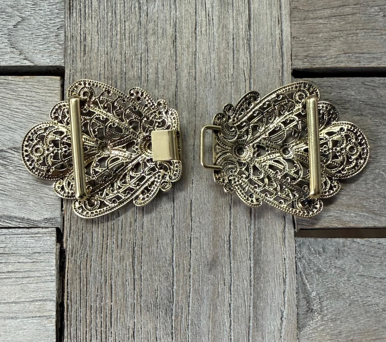 May include: Two-piece gold-colored filigree buckle set. Each piece has an ornate, openwork design with a bar and a clasp. The buckle is likely for a belt or other garment, and is displayed on a wooden surface.