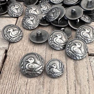 Puede incluir: Una colección de botones de metal plateados con un diseño en relieve de un pato en un estanque. Los botones son de forma ovalada y tienen un acabado brillante.