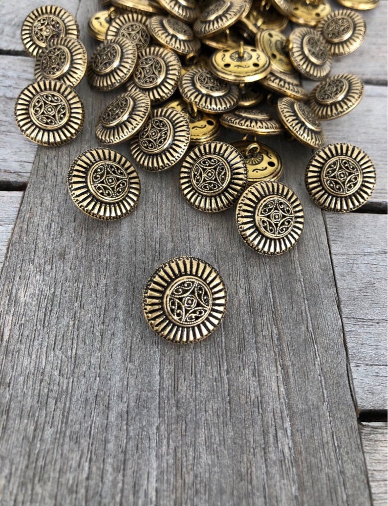 May include: A pile of gold-toned metal buttons with an ornate design. The buttons are round and have a raised center with a decorative pattern.