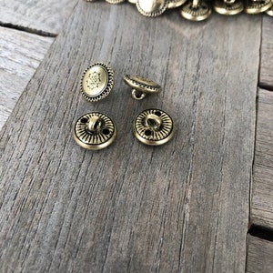 May include: Four gold-toned metal buttons with a decorative design. The buttons have two holes and a shank for sewing.