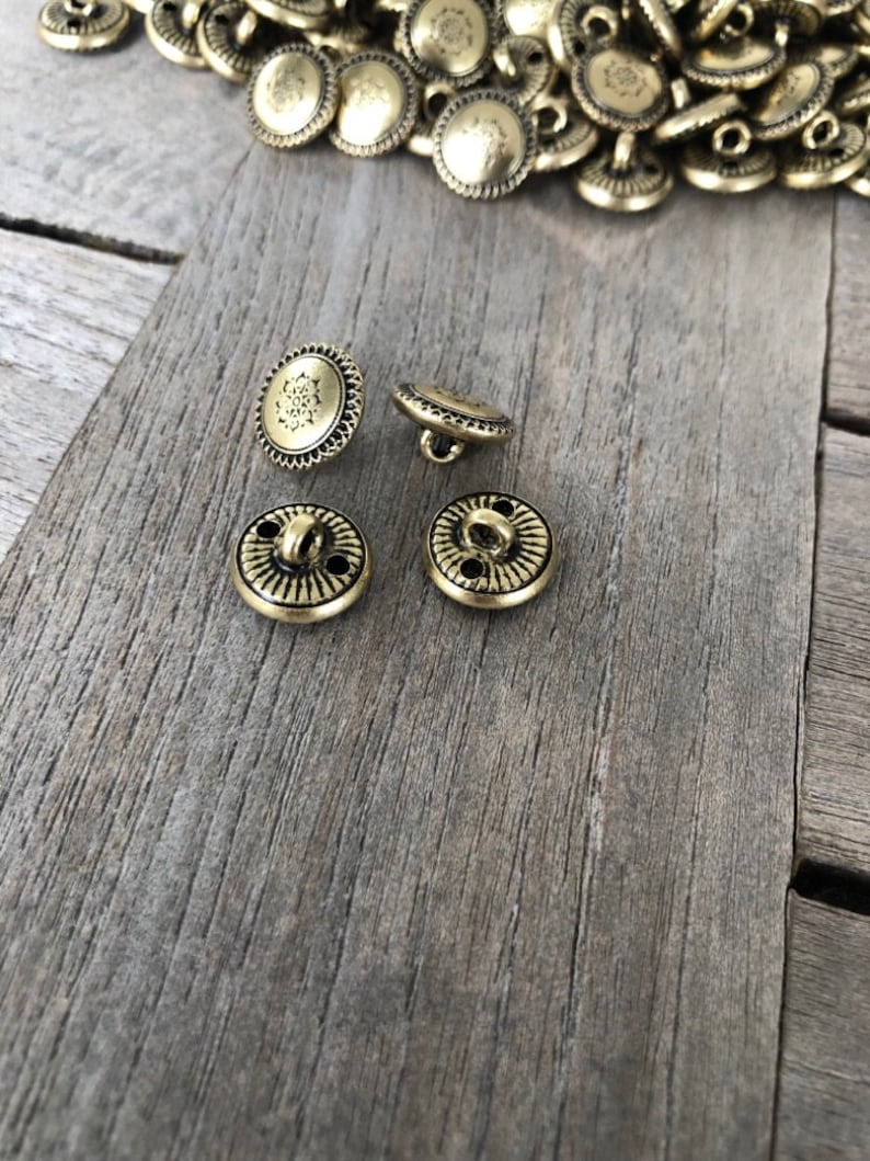 May include: Four gold-toned metal buttons with a decorative design. The buttons have two holes and a shank for sewing.
