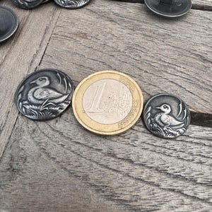 May include: A close-up of a silver metal button with a detailed design of a duck in a grassy area. The button is about the size of a US one-euro coin.