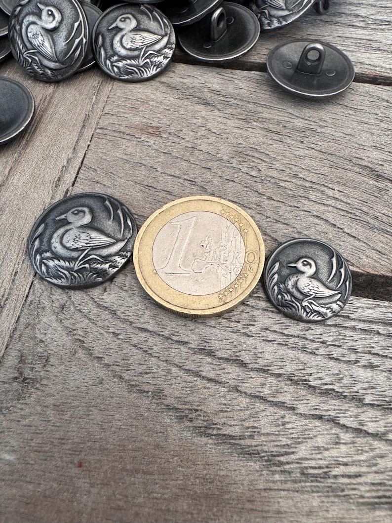 May include: A close-up of a silver metal button with a detailed design of a duck in a grassy area. The button is about the size of a US one-euro coin.