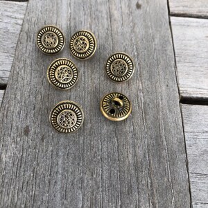 May include: Six gold-toned metal buttons with intricate designs. The buttons have a circular shape and a raised center.