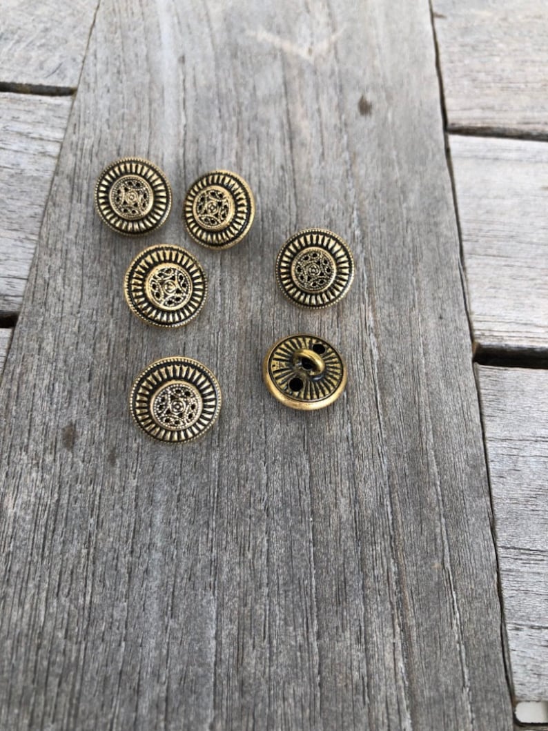 May include: Six gold-toned metal buttons with intricate designs. The buttons have a circular shape and a raised center.