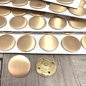 May include: Close-up of several sheets of gold-colored, round metal buttons. Some buttons are loose, while others are still attached to the white backing. One button has a decorative design on the back.