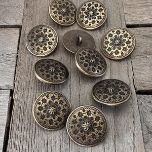 Könnte beinhalten: Eine Sammlung antiker Messingknöpfe mit floralem Design. Die runden Knöpfe haben ein erhabenes Blumenmuster und ein zentrales Design. Sie sind auf einer Holzoberfläche angeordnet.