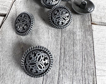 Pode incluir: Uma coleção de botões de metal prateados com designs florais intrincados. Os botões estão dispostos sobre uma superfície de madeira desgastada.