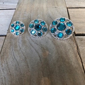 May include: Three round teal blue rhinestone buttons with silver metal frames. The buttons are arranged in order of size from smallest to largest.