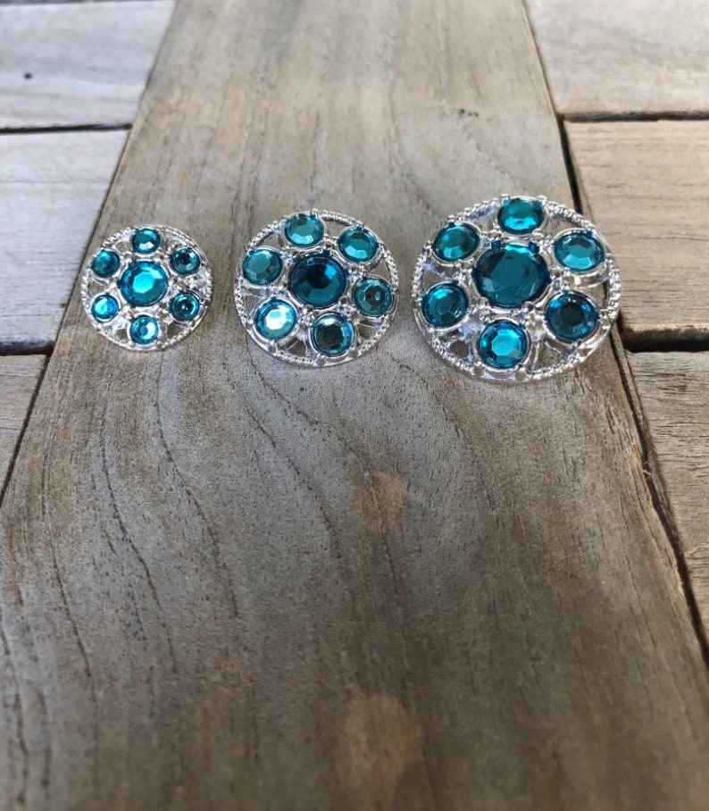 May include: Three round teal blue rhinestone buttons with silver metal frames. The buttons are arranged in order of size from smallest to largest.