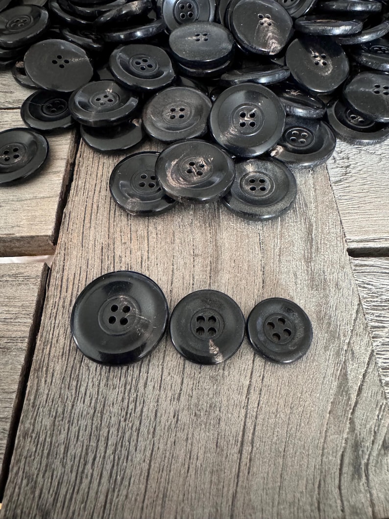 May include: A collection of black plastic buttons with four holes each. The buttons are arranged in a pile and on a wooden surface.