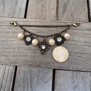 K&ouml;nnte beinhalten: Ein antikes Schmuckst&uuml;ck in Bronzet&ouml;nen mit Kette und Verschluss. Es verf&uuml;gt &uuml;ber Kunstperlen, klare Kristalle und verzierte Filigranmuster. Das Schmuckst&uuml;ck ist etwa 2.5 cm lang, mit einer M&uuml;nze zum Gr&ouml;&szlig;envergleich.