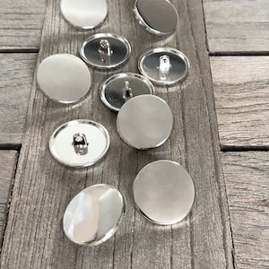 Pode incluir: Um grupo de dez botões redondos prateados com uma superfície plana e um eixo na parte de trás.