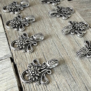 Puede incluir: Cierres metálicos plateados con diseño floral. Los cierres tienen una apariencia detallada y ornamentada con una flor central y detalles en espiral. Los cierres están dispuestos sobre una superficie de madera.