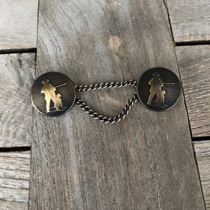 Könnte beinhalten: Ein Paar antike goldfarbene Metall-Hemdknöpfe mit einem Kettenverbinder. Jeder Knopf zeigt eine Silhouette einer Person mit einem Hund.