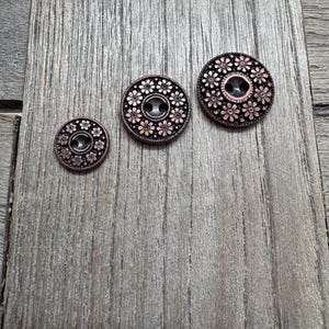 May include: Three copper-colored buttons with a floral design. The buttons are arranged from small to large, each featuring a central hole and a pattern of small flowers around the edge. The buttons are on a wooden surface.