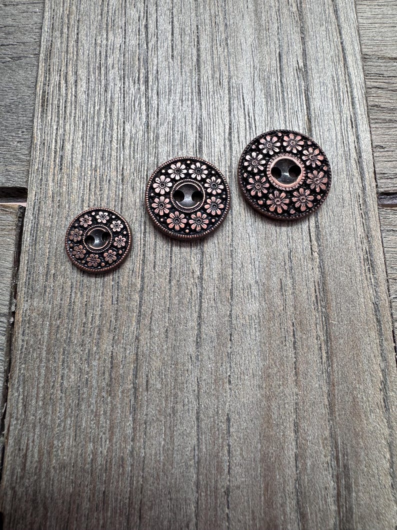 May include: Three copper-colored buttons with a floral design. The buttons are arranged from small to large, each featuring a central hole and a pattern of small flowers around the edge. The buttons are on a wooden surface.