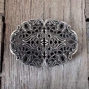 Könnte beinhalten: Verzierte silberfarbene Gürtelschnalle mit aufwendigem filigranem Design. Die Schnalle weist symmetrische Wirbelmuster und florale Motive auf. Die Metallarbeiten sind detailliert und haben eine Vintage- oder Antik-Ästhetik. Die Schnalle wird auf einer verwitterten Holzoberfläche präsentiert.
