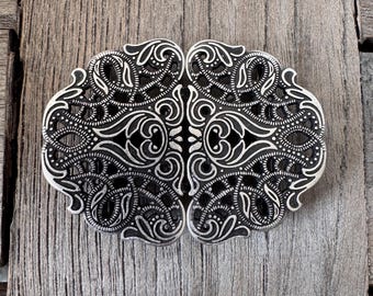 Pode incluir: Fivela de cinto prateada ornamentada com um intrincado design vazado. A fivela apresenta padrões de redemoinhos simétricos e motivos florais. O trabalho em metal é detalhado e tem uma estética vintage ou antiga. A fivela é exibida em uma superfície de madeira desgastada.