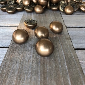 Op de afbeelding: Een groep gouden knoppen met een glanzende afwerking. De knoppen zijn rond en hebben een platte achterkant. Er zijn verschillende knoppen verspreid over een houten oppervlak, en een paar meer zijn in een lijn gerangschikt.