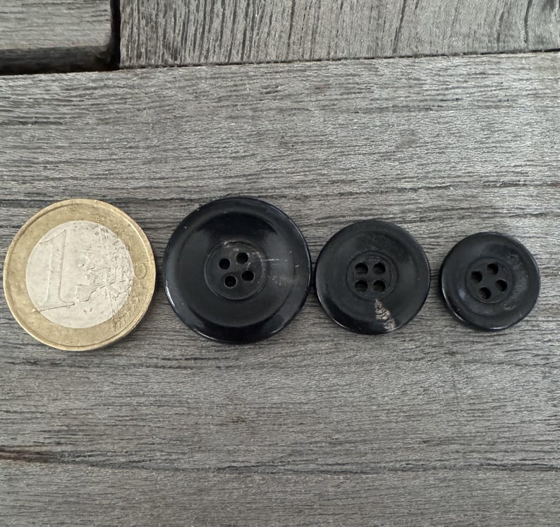 May include: Three black plastic buttons with four holes each. The buttons are arranged in a row from largest to smallest.