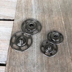 May include: A set of four black metal snap fasteners. The fasteners are in two sizes, with two larger fasteners and two smaller fasteners. The fasteners are on a wooden surface.