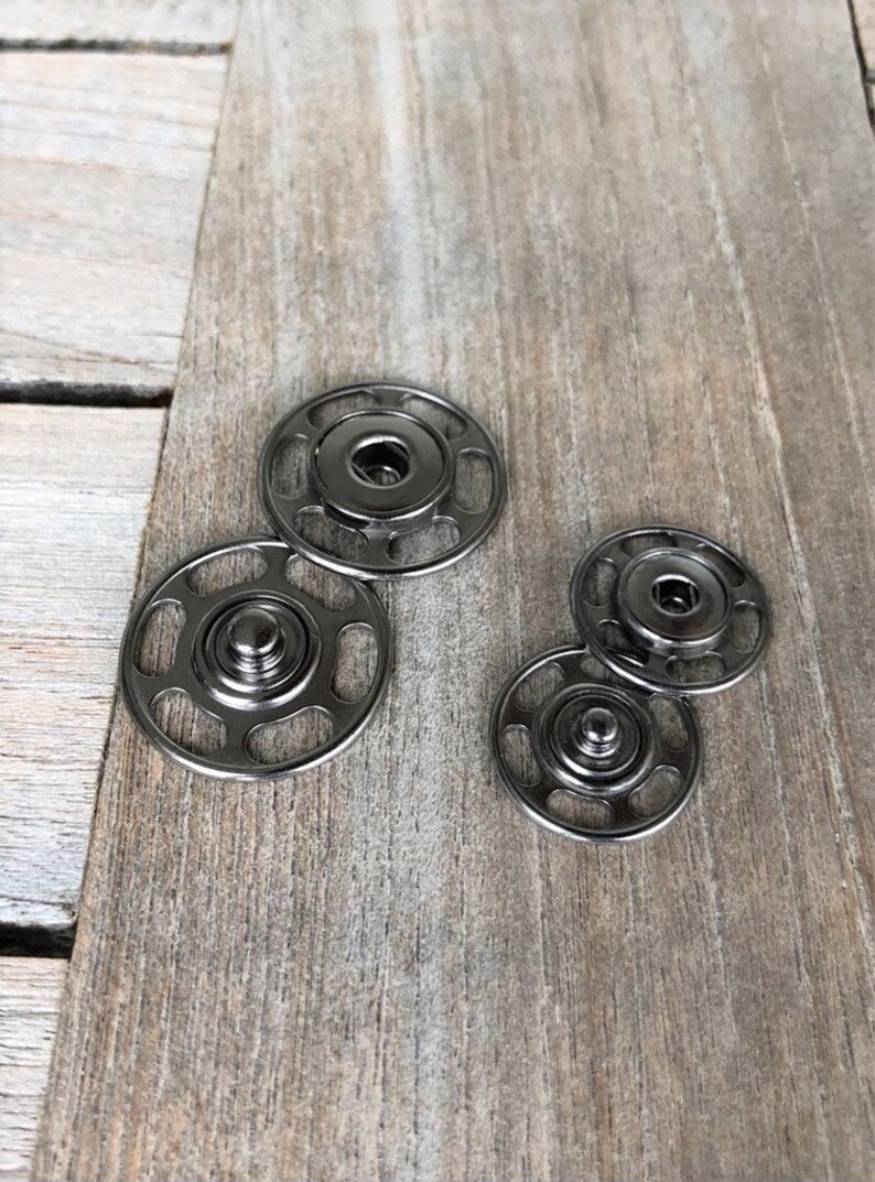 May include: A set of four black metal snap fasteners. The fasteners are in two sizes, with two larger fasteners and two smaller fasteners. The fasteners are on a wooden surface.