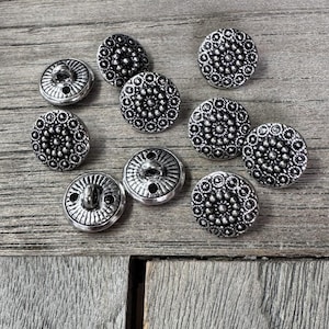 Peut inclure: Un assortiment de boutons argentés avec des motifs floraux complexes. Les boutons sont ronds avec un motif détaillé de petits cercles et un trou central pour la couture. Ils sont disposés sur une surface en bois vieilli.