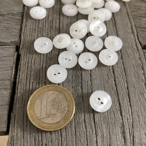 May include: A pile of white plastic buttons with two holes each. The buttons are scattered on a wooden surface next to a Euro coin.