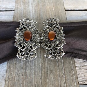 Puede incluir: Un par de hebillas de cintur&oacute;n de metal plateado con dise&ntilde;os florales ornamentados y piedras de vidrio color &aacute;mbar.