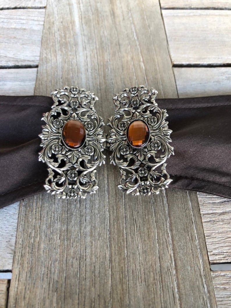 Puede incluir: Un par de hebillas de cintur&oacute;n de metal plateado con dise&ntilde;os florales ornamentados y piedras de vidrio color &aacute;mbar.