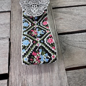 K&ouml;nnte beinhalten: Eine silberne G&uuml;rtelschnalle mit einem komplizierten Design, befestigt an einem gewebten G&uuml;rtel mit schwarzem Hintergrund und einem bunten Blumenmuster.
