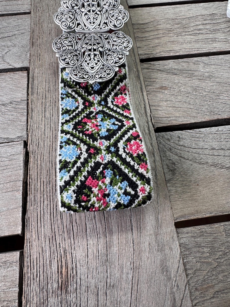K&ouml;nnte beinhalten: Eine silberne G&uuml;rtelschnalle mit einem komplizierten Design, befestigt an einem gewebten G&uuml;rtel mit schwarzem Hintergrund und einem bunten Blumenmuster.