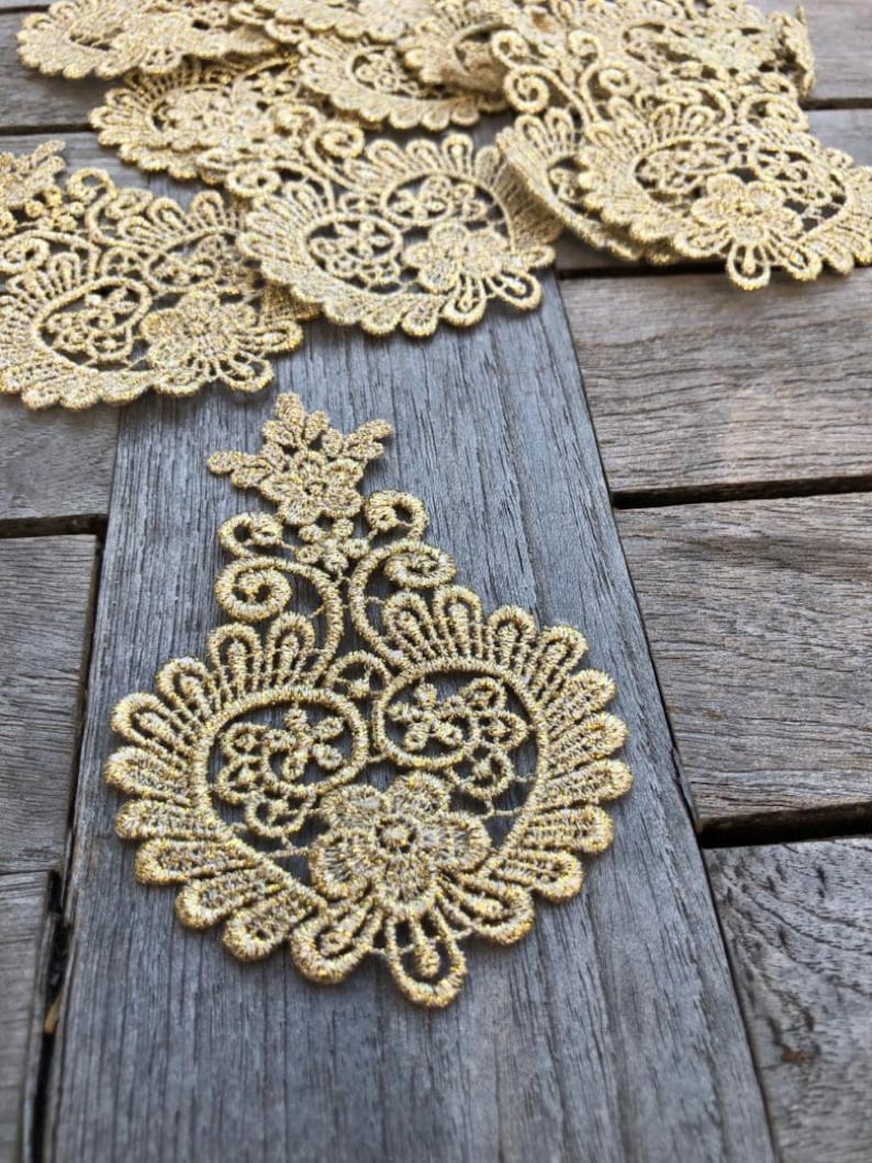 May include: Gold lace trim with a delicate floral pattern. The lace is arranged in a decorative pattern on a wooden surface.