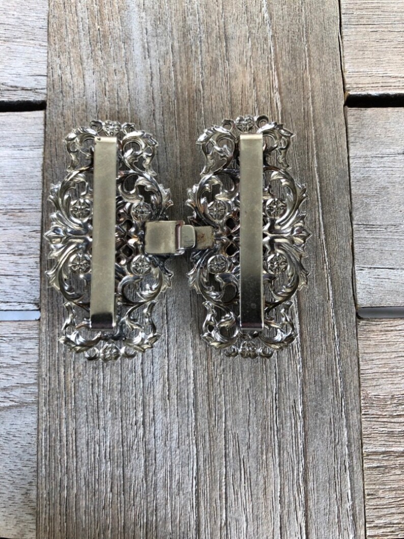 Puede incluir: Un par de hebillas de cintur&oacute;n de metal plateadas con intrincados dise&ntilde;os florales. Las hebillas est&aacute;n conectadas por una barra de metal.