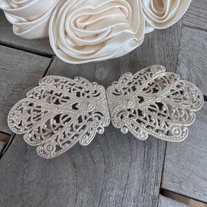 K&ouml;nnte beinhalten: Eine silberne Filigran-G&uuml;rtelschnalle mit einem zarten, filigranen Design. Die Schnalle ist aus Metall und hat eine gl&auml;nzende Oberfl&auml;che.
