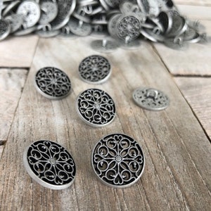 Puede incluir: Un montón de botones de metal plateado con un diseño de filigrana negra. Los botones están dispuestos en una pila sobre una superficie de madera.