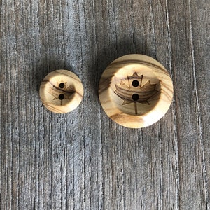 May include: Two wooden buttons with a maple leaf design. The larger button has a more detailed design and the smaller button is plain.