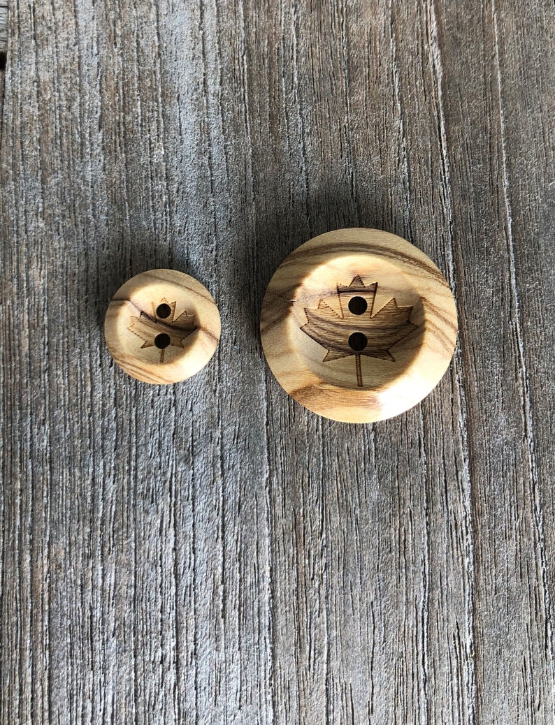 May include: Two wooden buttons with a maple leaf design. The larger button has a more detailed design and the smaller button is plain.