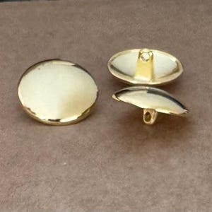 Könnte beinhalten: Drei goldfarbene Knöpfe. Einer ist rund, und zwei sind oval mit einer Schlaufe auf der Rückseite. Die Knöpfe haben eine glänzende, reflektierende Oberfläche. Die Knöpfe liegen auf einer braunen Oberfläche.