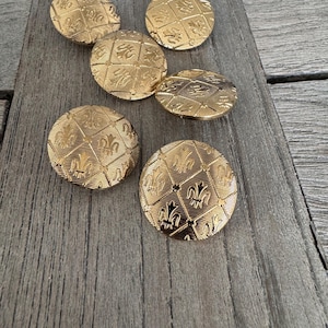 Könnte beinhalten: Sechs goldfarbene Knöpfe mit einem dekorativen Design, das ein sich wiederholendes Muster aus Fleur-de-lis und Diamanten zeigt. Die Knöpfe sind auf einer Holzoberfläche angeordnet.