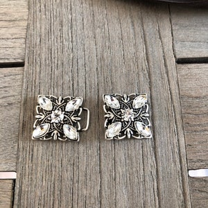 Może przedstawiać: Dwie srebrne klamry do paska z przezroczystymi kryształkami. Każda klamra ma kwadratowy kształt z ozdobnym wzorem kwiatowym.