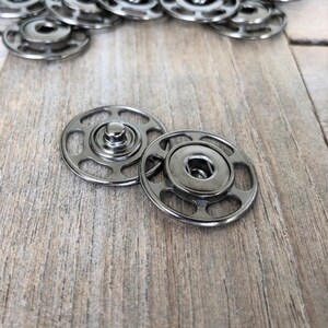 May include: Two silver metal snap fasteners, one on top of the other, on a light brown wooden surface.