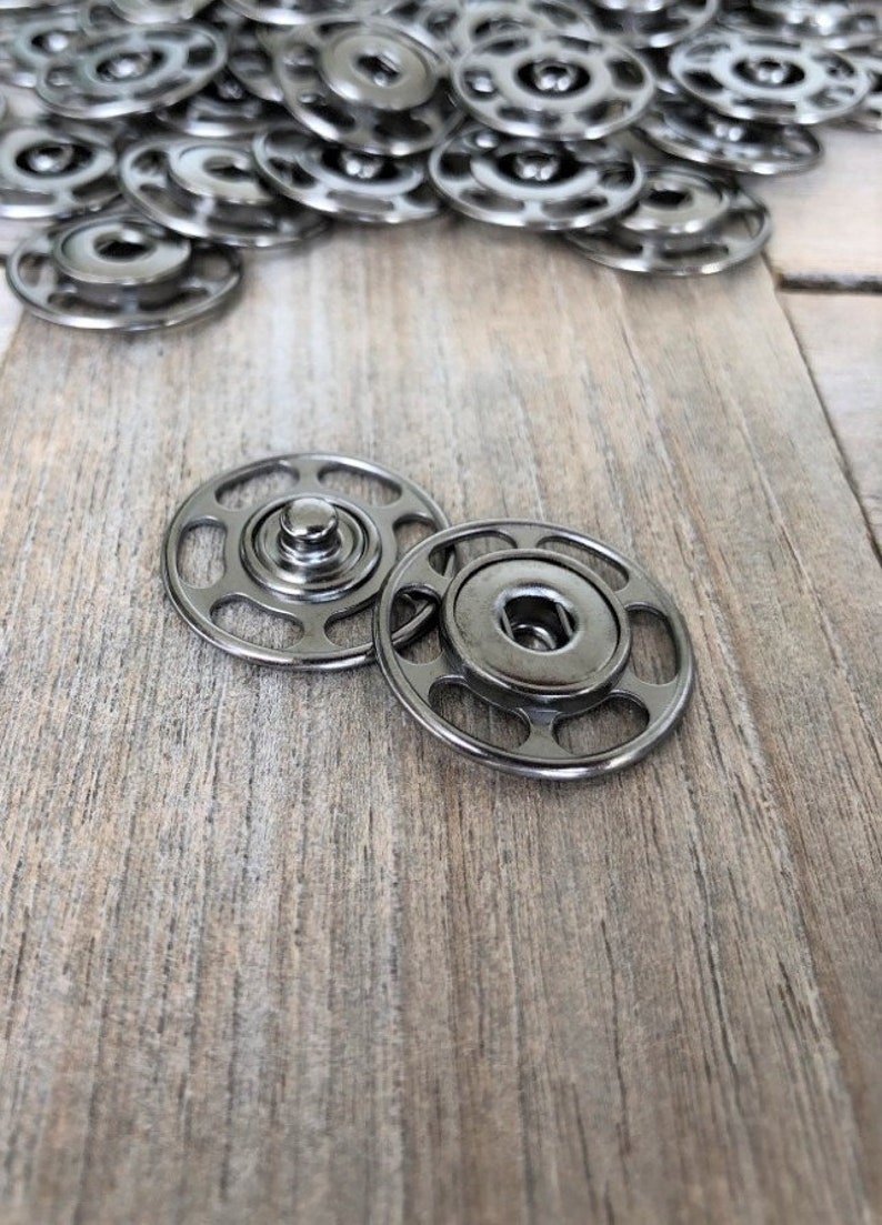 May include: Two silver metal snap fasteners, one on top of the other, on a light brown wooden surface.