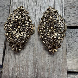 May include: Two ornate, antique-style brass clasps with intricate floral and scrollwork designs. The clasps are oval-shaped and feature a detailed, symmetrical pattern. They are set against a weathered, wooden background.