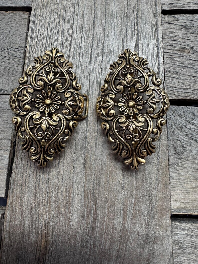May include: Two ornate, antique-style brass clasps with intricate floral and scrollwork designs. The clasps are oval-shaped and feature a detailed, symmetrical pattern. They are set against a weathered, wooden background.