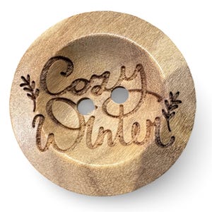 May include: A round wooden button with two holes and the words "Cozy Winter" engraved in a cursive font. The button has a light brown color with a natural wood grain texture. Small leaf designs flank the text.