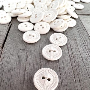 Pode incluir: Um monte de botões de cerâmica branca com um design em relevo. Os botões estão dispostos em um monte sobre uma superfície de madeira. Há cinco botões em primeiro plano, com um design mais intrincado.