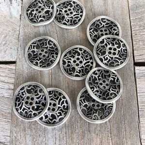 Może przedstawiać: Zestaw 10 srebrzystych metalowych guzików z ozdobnym wzorem filigranowym. Guziki mają okrągły kształt i posiadają wypukłe centrum z pięcioma otworami.